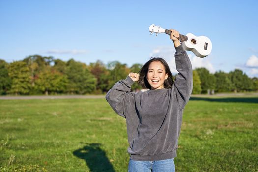 Upbeat young woman dancing with her musical instrument. Girl raises her ukulele up and pose in park on green field