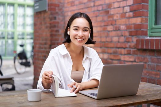 Stylish asian girl works in coworking with laptop, makes notes, uses wireless earphones and smiles, sits near brick wall in office