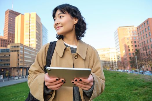 Happy young brunette girl, asian woman walks around city with tablet, goes to university with her digital gadget and backpack
