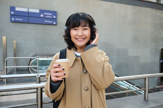 Beautiful smiling Korean girl, drinks takeaway coffee, listens music in headphones, wears trench and backpack, stands on train station, travelling