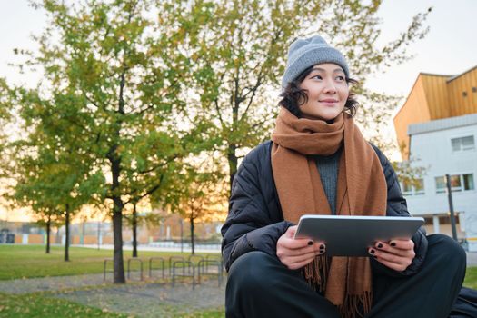 Portrait of asian girl in warm clothes, sits on bench with digital tablet and graphic pen, smiling happily, draws outdoors in chilly weather