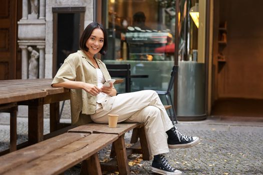 Portrait of stylish young korean woman sits in cafe, holds smartphone, smiles, enjoys coffee outdoors