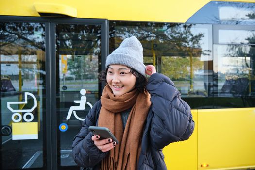 Portrait of girl standing near bus on a stop, waiting for her public transport, schecks schedule on smartphone application, holds mobile phone, wears warm clothes