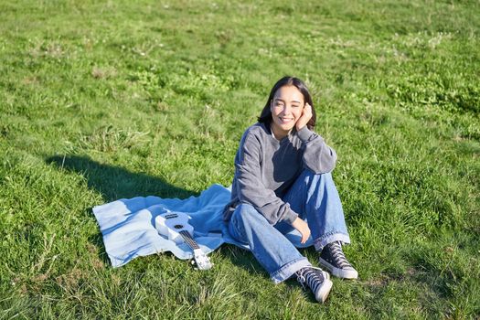 Young asian girl enjoying sunny day outdoors. Happy student having picnic on grass in park, playing ukulele
