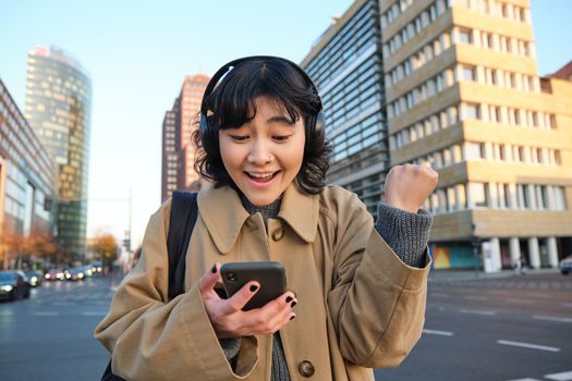Young happy woman celebrating on street, holding smartphone and cheering, reacts amazed to good news, reads phone text message with surprised joyful face