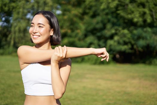 Portrait of young fitness woman stretching her arms, warm-up before training session, sport event in park, jogging and excercising.