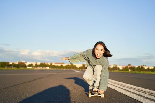 Skater girl riding on skateboard, standing on her longboard and laughing, riding cruiser on an empty street towards the sun