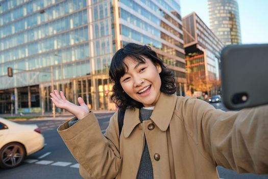 Smiling korean girl showing city, taking selfie in front of buildings on street with happy face, takes photos on her smartphone camera