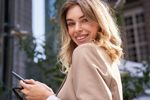 Close up portrait of successful young woman, corporate lady in suit, holds her mobile phone, turns at camera with happy smile, stands on street in daytime