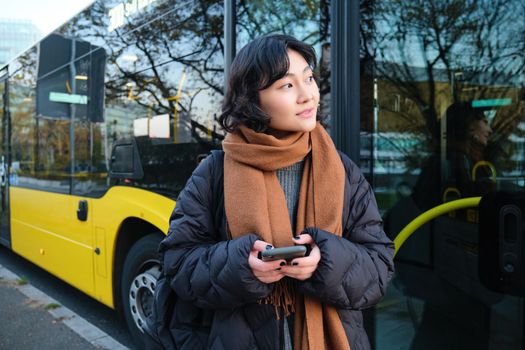 Portrait of korean girl buying ticket for public transport online, using mobile application on bus stop, wearing winter clothes.