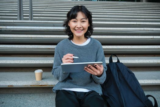 Portrait of asian girl student, hipster sitting on stairs with digital tablet and cup of coffee, draws digital art, makes design project