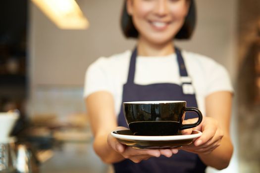 Cropped shot, female barista hands holding cup of coffee, giving you drink, serving in cafe, wearing uniform