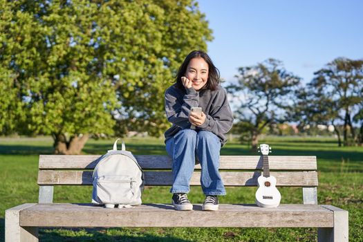 Beautiful brunette girl on bench in park, sitting with ukulele and backpack, holding smartphone, using mobile app