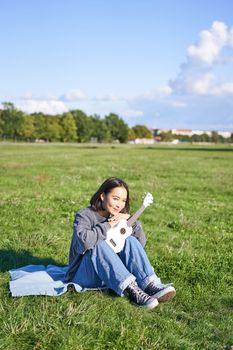 Portrait of asian girl sitting alone in park, playing ukulele and singing, enjoying leisure time
