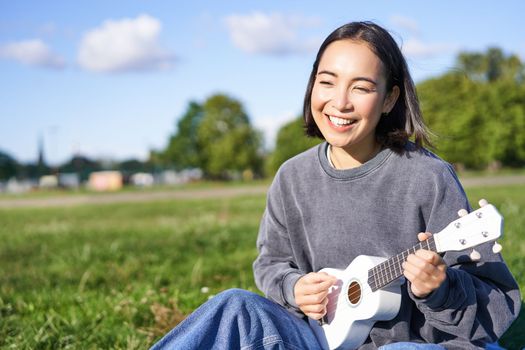 Carefree asian girl singing and playing ukulele in park, sitting on grass, musician relaxing on her free time outdoors
