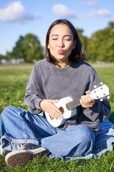 Positive hipster asian girl, playing ukulele and making faces, singing and feeling happy