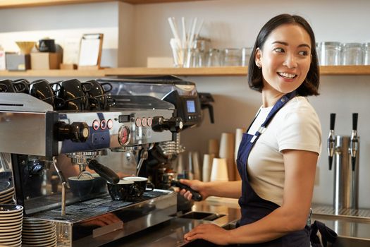 Smiling asian barista girl makes cappuccino with coffee machine, stands behind counter in cafe