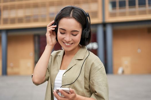 Streaming services concept. Happy asian girl listens music in headphones, holds mobile phone, choosing track or podcast, walking on street