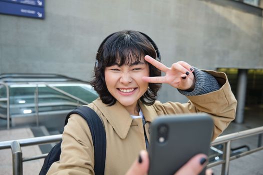 Beautiful korean girl in headphones, takes selfie on her smartphone, stands on street with mobile phone, records video, listens music and travels around city