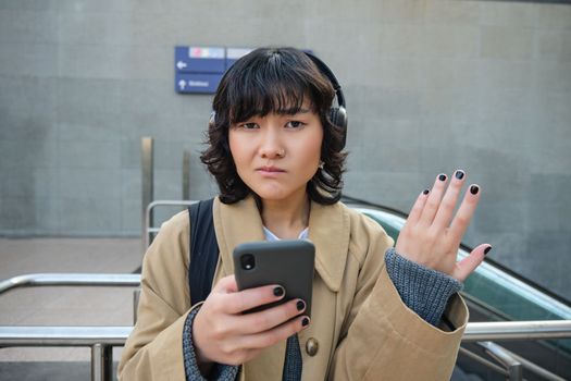 Portrait of asian girl in headphones, looks complicated at smartphone screen, puzzled by text message or notification, stands on street and shrugs