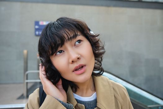 Portrait of stylish asian girl in headphones, listens podcast or music with confused face, looking up puzzled, seeing smth strange, standing on street