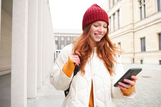 Beautiful smiling girl, tourist with backpack, holding smartphone, using map on mobile phone application, looking for sightseeing in internet app, standing outdoors