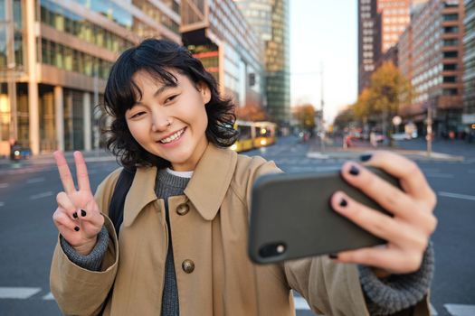Happy asian girl, tourist takes photo in city centre, shows peace sign at smartphone camera, makes selfie during sightseeing with smiling joyful face