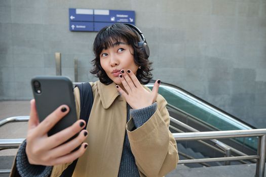 Cute and funny korean girl in headphones, posing for selfie, takes photo on smartphone and looks silly, stands on street in city