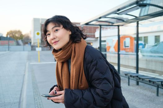 Cute smiling woman standing on a bus stop and looking at road, waiting for her public transport, holding smartphone, checking schedule on mobile app
