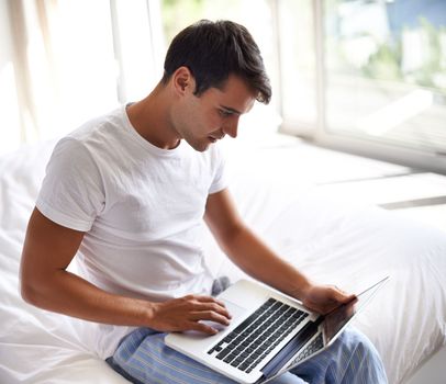 Checking his emails in the AM. a handsome young man using a laptop on the edge of his bed.