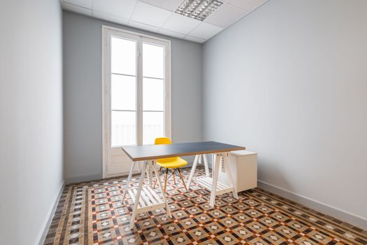 An empty room with gray smooth walls, doors leading to terrace. Floor is made of patterned marble tiles. There is table with gray top a yellow plastic chair with wooden legs in room.
