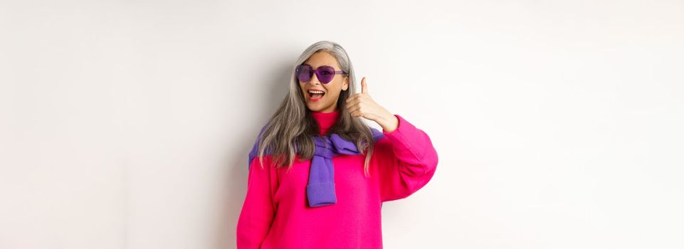 Shopping concept. Stylish asian senior woman in sunglasses and trendy outfit, showing thumb-up in approval, recommending shop, standing over white background