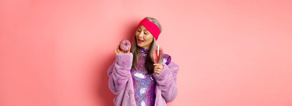 Fancy senior woman having fun, eating donut and drinking pink champagne, standing in purple faux fur coat and glittering dress, studio background