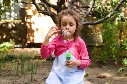 Adorable Caucasian little girl with beautiful curly long hair, blowing soap bubbles in the nature at sunset