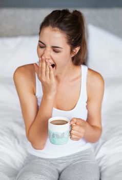 Still so sleepy...A young woman giving a big yawn while sitting in bed with a cup of coffee.