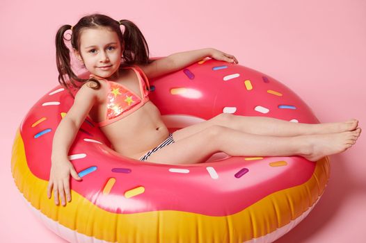 View from above of charming little child girl relaxing on a floating donut swim ring, isolated on pink color background