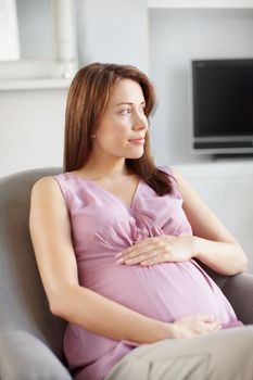 Experiencing the perfect pregnancy. Pretty young pregnant woman looking away while she sits in her living room as she holds her belly.