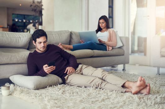I am so glad that we can relax the whole day. a young couple relaxing in the living room while being on their mobile devices at home.