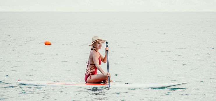 Woman in red bikini on sup board. Happy lady with blond hair in red bathing suit chilling and sunbathing by turquoise sea ocean on sunset. Holiday, vacation and recreational concept.