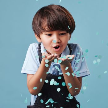 An adorable asian little boy having fun while blowing blue confetti at a gender reveal party. Cute mixed race child being curious and playful in a studio
