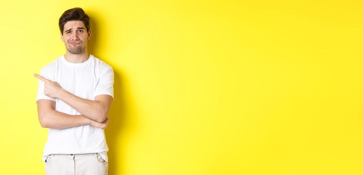 Skeptical and reluctant guy in white t-shirt, pointing finger left and smirking unamused, showing bad advertisement, standing over yellow background
