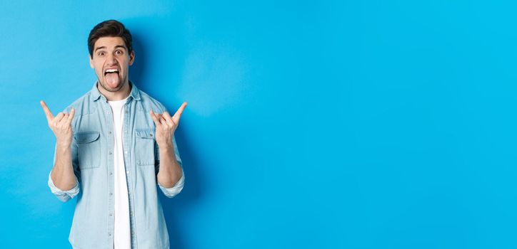 Excited handsome guy having fun, showing rock and roll sign and sticking tongue, standing against blue background