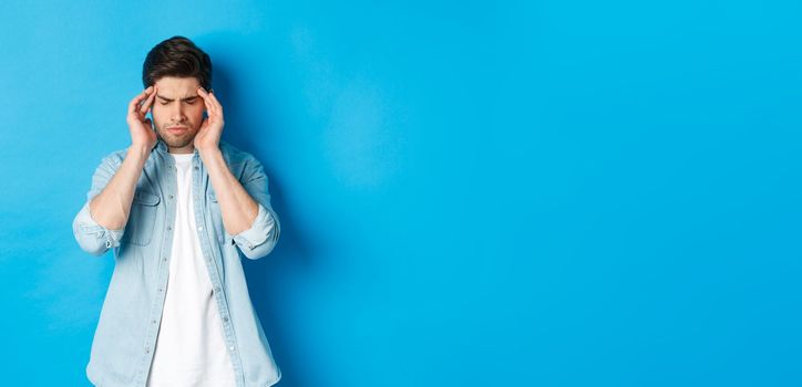 Man with hangover touching head and grimacing, having headache, standing against blue background