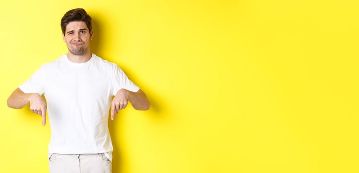 Young unhappy guy grimacing, pointing fingers down at advertisement, disappointed in product, standing over yellow background