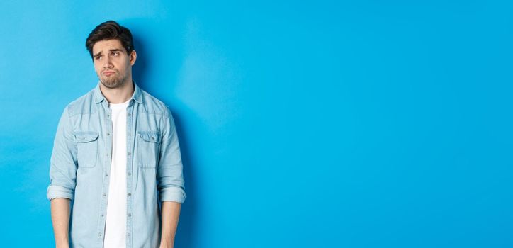 Image of reluctant and sad guy in casual outfit looking left, frowning and feeling upset, standing against blue background