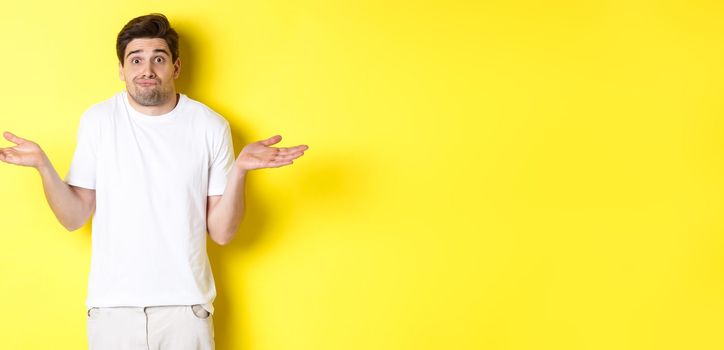 Clueless man shrugging, spread hands sideways confused, dont know anything, standing over yellow background
