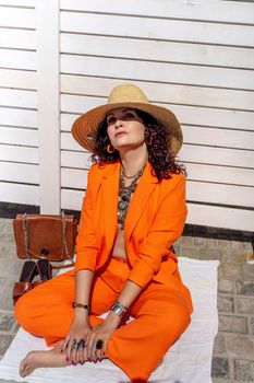 Stylish woman in an orange suit with a hat sits on a rug on a white striped background. On the hands are jewelry rings and bracelets, sandals and a bag stand side by side.