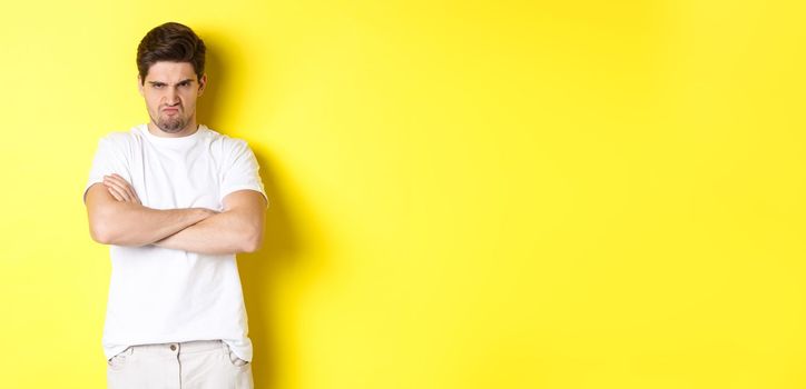 Offended and mad guy, frowning and sulking, holding hands crossed on chest, standing angry against yellow background