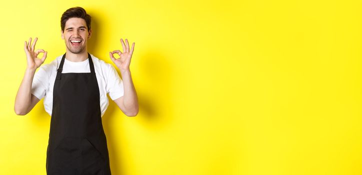 Young male barista in black apron showing OK signs and smiling, guarantee quality in coffee shop, standing over yellow background