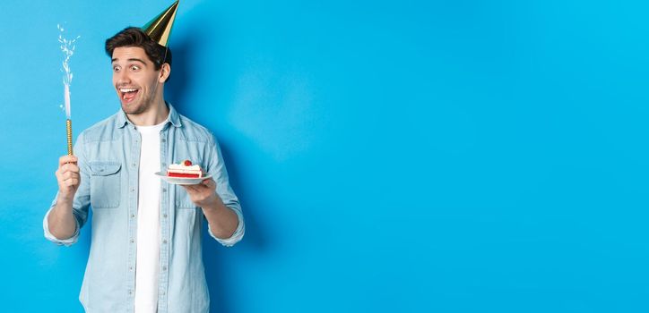 Happy young man celebrating birthday in party hat, holding b-day cake and smiling, standing over blue background